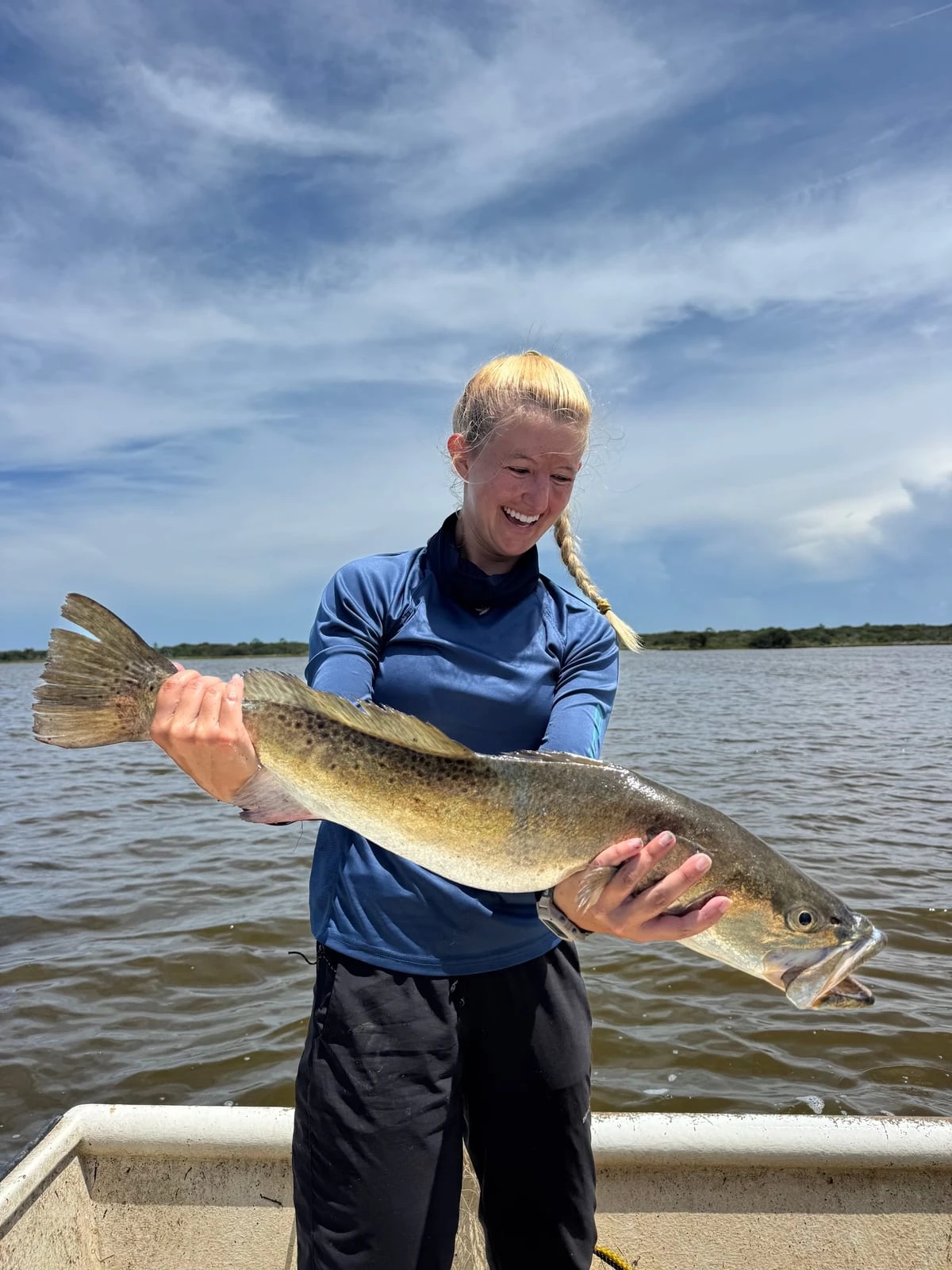 On a boat in a blue fishing shirt, holding a large speckled trout, with open water behind her