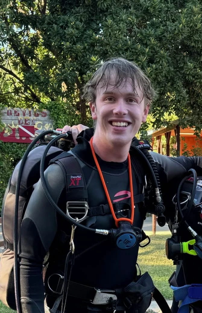 Smiling in a wetsuit and dive gear, hair still wet, in front of a leafy tree